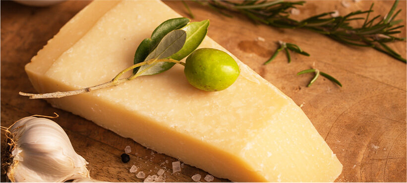 Parmigiano reggiano on a wood cutting board with fresh rosemary, garlic, and coarse salt with a green olive on top.